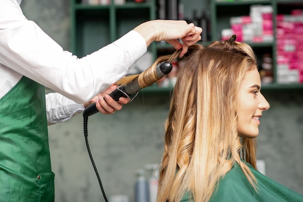 Criação de cachos com ferros de ondulação. cabeleireiro faz um penteado para uma jovem de cabelos vermelhos compridos em um salão de beleza.