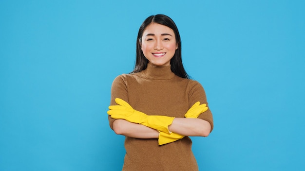 Criada profesional sonriente con guantes de goma protectores de pie con el brazo cruzado en el estudio sobre fondo azul. Alegre ama de llaves asiática tuvo mucho cuidado en las tareas de limpieza de la casa del cliente