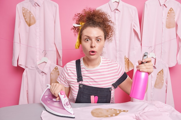 La criada responsable de la mujer de pelo rizado mira fijamente los ojos saltones ocupados con las tareas del hogar se encuentra cerca de la mesa de planchado sostiene una botella de spray de agua y soportes de plancha eléctrica cerca de las camisas planchadas. Concepto de limpieza