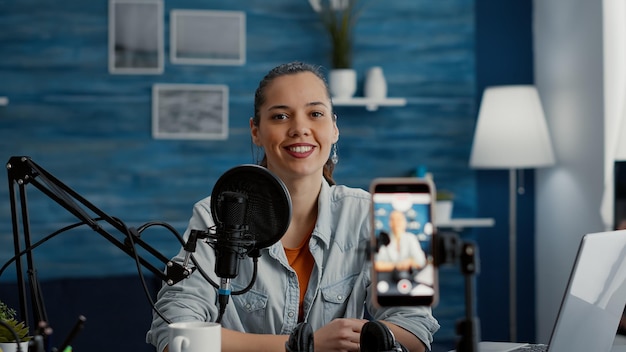 Criador de conteúdo digital criativo sentado na mesa do home studio enquanto filmava vlog com smartphone. Feliz e sorridente jovem influenciador de mídia social se preparando para gravar um blog de vídeo enquanto está no estúdio.