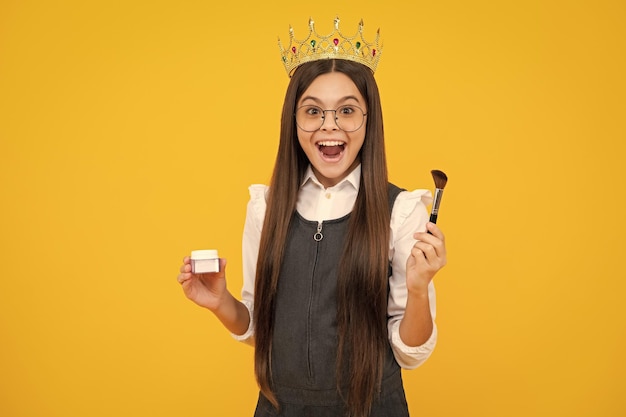 Criança adolescente excitada na coroa da rainha segura pó e escova para maquiagem de aniversário em amarelo