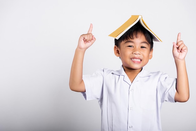 Criança adorável asiática sorrindo feliz vestindo uniforme tailandês de estudante segurando livro sobre a cabeça como telhado