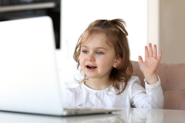 Criança adorável sorridente usa um chat de vídeo de laptop para se comunicar enquanto está sentado em uma mesa em casa. Garoto online.