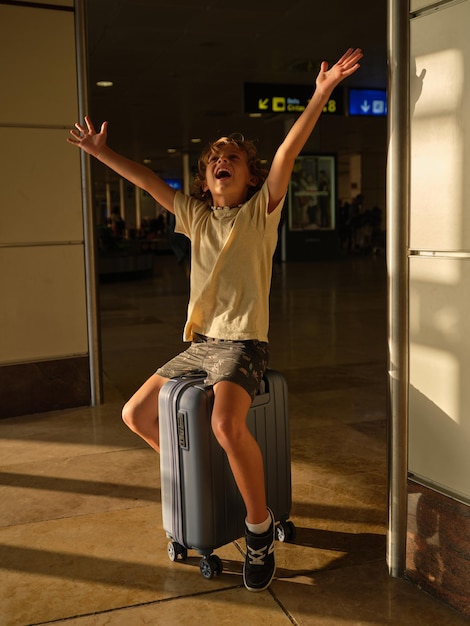 Foto criança alegre com os braços levantados sentado na bagagem e esperando a partida no aeroporto enquanto viaja durante as férias de verão