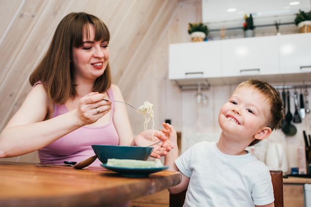 Criança alegre comendo macarrão na cozinha com a mãe