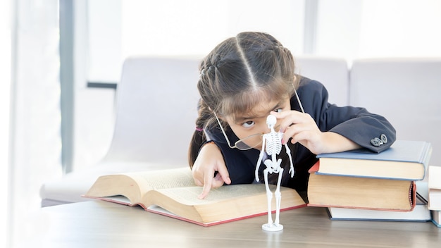 Criança asiática aprendendo ciências biológicas em livro com modelo de esqueleto