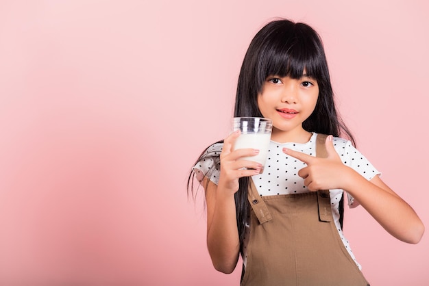 Criança asiática de 10 anos sorri segura copo de leite bebe leite branco e aponta o dedo