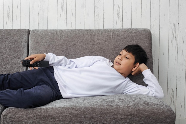 Criança asiática de suéter branco está relaxando no sofá com expressão de sorriso enquanto assiste televisão