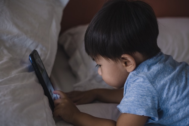 Criança asiática, deitada na cama e brincando no celular