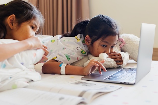 Criança asiática doente se sente entediada e deitada na cama com sua irmã enquanto aprende uma aula online com um professor e um amigo quando está no hospital. Ensino doméstico de isolamento e ensino à distância.