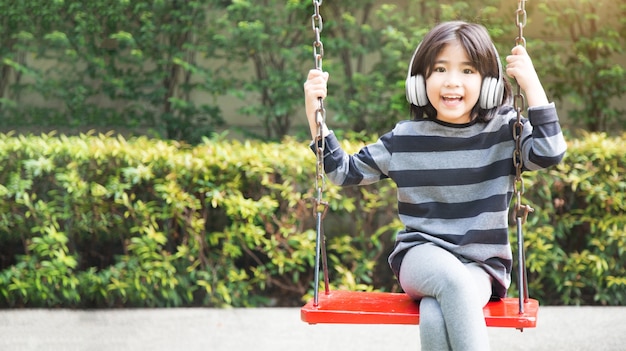 Criança asiática feliz ouvindo música com fone de ouvido sem fio e jogando swing no parque kidtergarden com estilo de vida digital na horizontal
