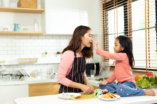 Criança asiática pré-escolar faz bolo cozinhando na cozinha Mãe prepara decoração bonita com diversão educa cupcake para irmã de aniversário Creme bagunça em todo o cabelo rosto boca sinta feliz espaço de cópia