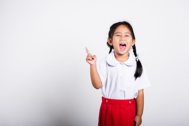 Criança asiática sorrindo feliz vestindo saia vermelha uniforme tailandesa de estudante continua apontando o dedo para o espaço da cópia