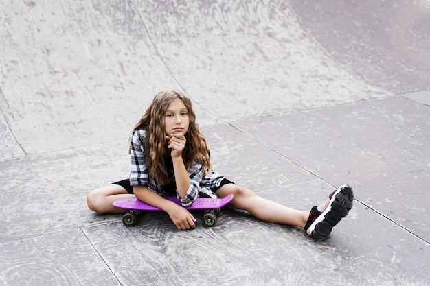 Criança atraente com penny board sentado na rampa esportiva no playground de skate Adolescente ativo posando com penny board Estilo de vida extremo