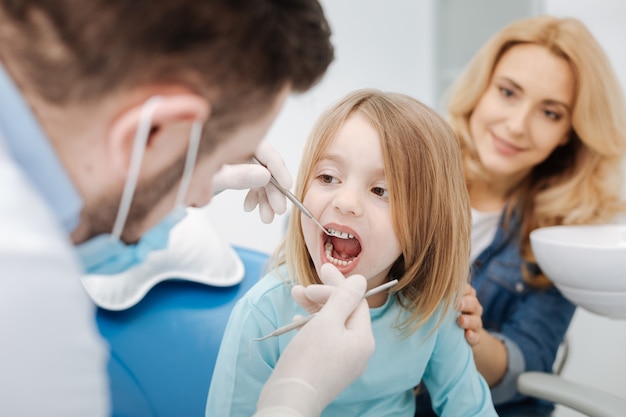 Criança boa e paciente de bom temperamento visitando seu médico regularmente e sentada quieta com a boca aberta enquanto ele examina seus dentes