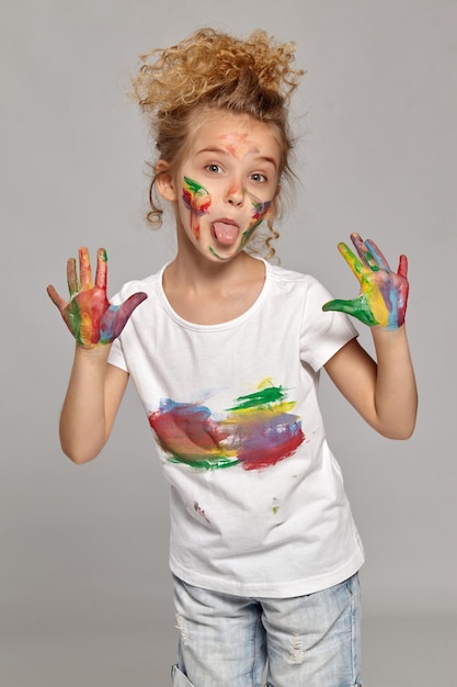 Criança bonita com uma escova em seu lindo corte de cabelo, vestindo uma camiseta branca manchada. Ela está posando com as mãos e bochechas pintadas e mostrando a língua, em um fundo cinza.
