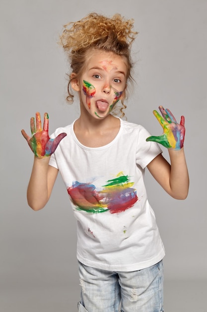 Foto criança bonita com uma escova em seu lindo corte de cabelo, vestindo uma camiseta branca manchada. ela está posando com as mãos e bochechas pintadas e mostrando a língua, em um fundo cinza.