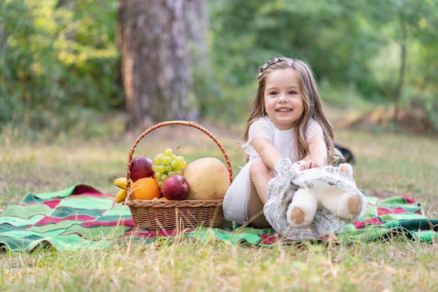 Criança bonita no piquenique com cesta Criança descansando no parque ou jardim ensolarado Menina com ursinho e aproveitando o lazer