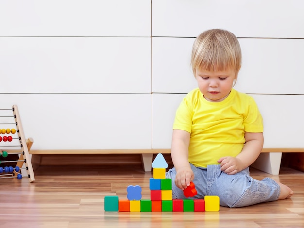 Criança bonitinha em uma camiseta amarela está brincando com cubos de brinquedo no chão da casa