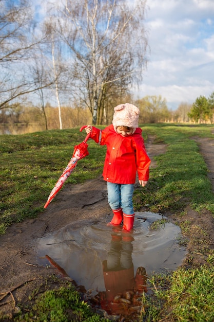 Criança brinca com guarda-chuva depois da chuva em botas de borracha vermelha e capa de chuva
