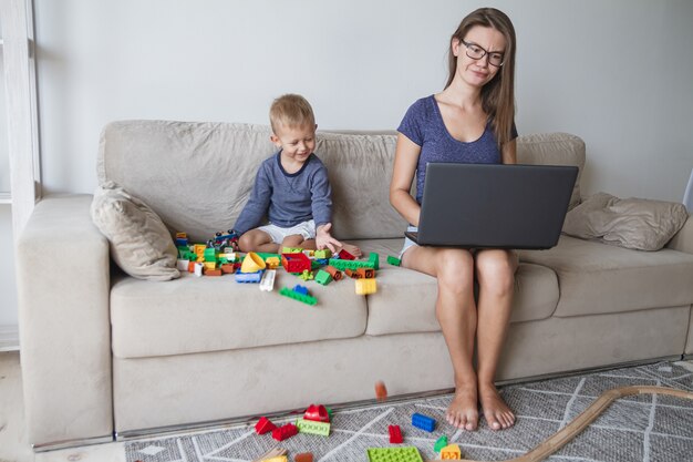 Criança brincando no sofá enquanto a mãe trabalha no laptop