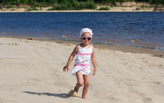 Criança caucasiana caprichosa e infeliz de três anos em óculos de sol andando na praia do rio de areia