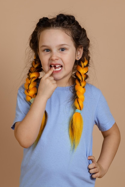 Foto criança caucasiana pequena com o dedo apontando para a boca mostrando falta de dente que dói olhando para a câmera com tranças kanekalon na cabeça no fundo bege em camiseta azul claro