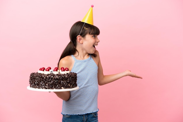 Criança caucasiana segurando bolo de aniversário isolado em fundo rosa com expressão de surpresa enquanto olha de lado