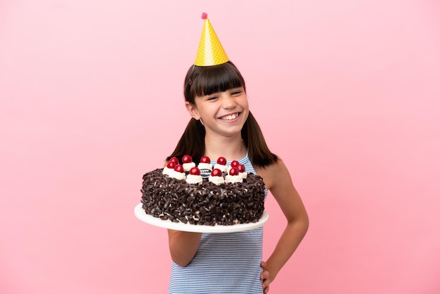 Criança caucasiana segurando bolo de aniversário isolado em fundo rosa posando com braços no quadril e sorrindo