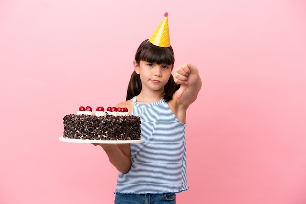 Criança caucasiana segurando o bolo de aniversário isolado no fundo rosa, mostrando o polegar para baixo com expressão negativa