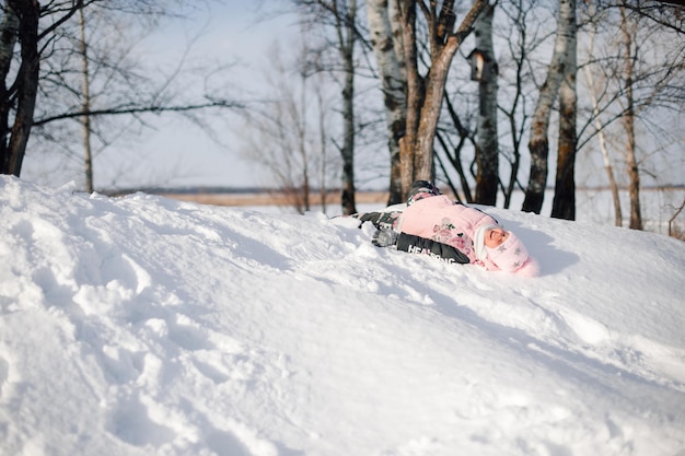 Criança cavalga no escorregador de neve menina gosta de caminhada de inverno na natureza na floresta, brinca de brincadeira e deita na neve