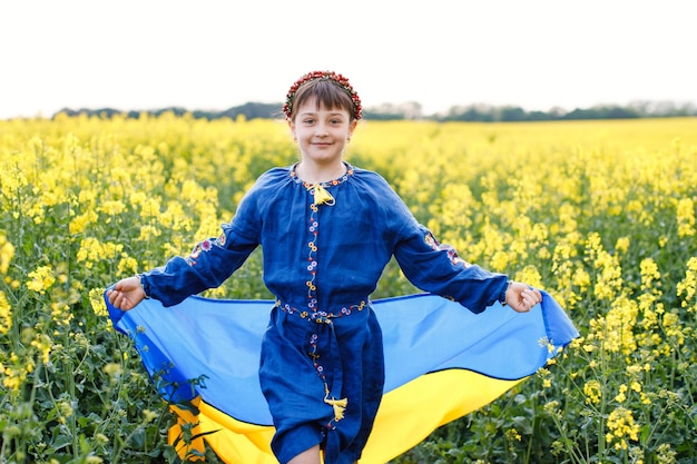 Criança com bandeira ucraniana em campo de colza Uma menina de camisa bordada atravessa o campo com a bandeira ucraniana nas mãos