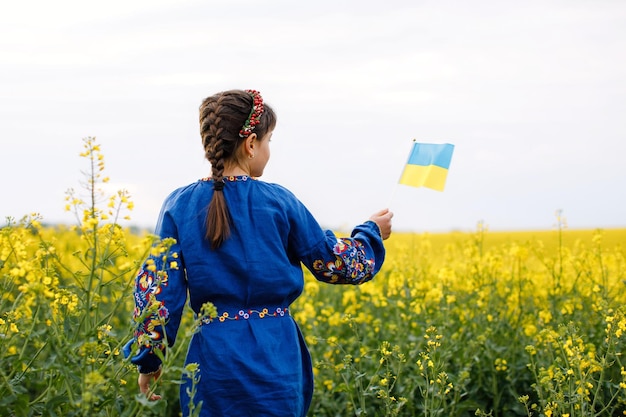 Criança com bandeira ucraniana em campo de colza Uma menina de camisa bordada atravessa o campo com a bandeira ucraniana nas mãos