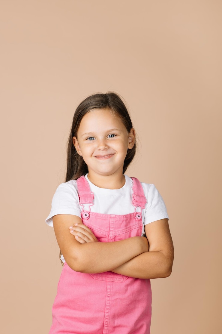 Foto criança com braços cruzados com olhos brilhantes e sorriso humilde olhando para a câmera vestindo macacão rosa brilhante e camiseta branca sobre fundo bege