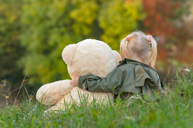 Criança com brinquedo macio descansa na natureza Menina loira e ursinho de pelúcia no fundo das árvores de outono Vista traseira