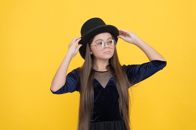 Criança com cabelo longo moreno consertar o chapéu preto com fundo amarelo da aparência da festa da moda, beleza.