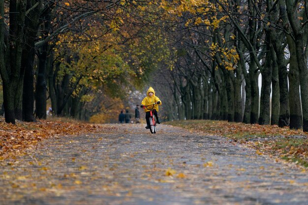 Criança com capa de chuva amarela anda de bicicleta no parque chuvoso de outono. Garoto solitário anda de bicicleta ao longo do beco de outono.