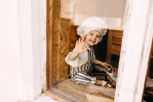 Criança com chapéu de chef e avental perto da janela sorrindo e acenando