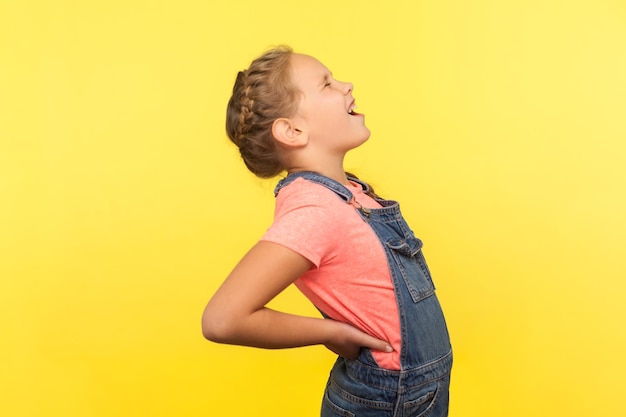 Criança com dor nas costas Retrato de menina insalubre desesperada em macacão jeans tocando a parte inferior da coluna e gritando de dor problema médico tiro de estúdio interno isolado em fundo amarelo