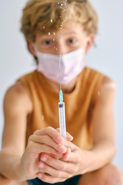 Criança com máscara facial olhando para a câmera enquanto borrifa a vacina do injetor durante a pandemia de COVID 19 em fundo claro