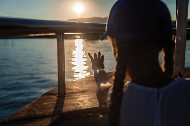 Foto criança com um boné azul ao pôr-do-sol em um barco tocando o reflexo da luz solar na água evocando uma sensação de