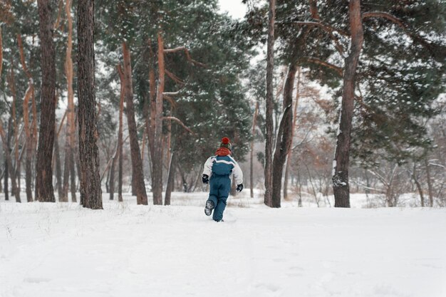 Criança corre em direção a grandes pinheiros de neve Menino brinca ao ar livre na floresta de inverno Vista traseira