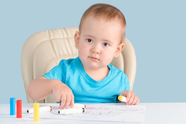 Criança de 1217 meses aprende a desenhar com canetas de feltro em um pedaço de papel sentado em uma cadeira fundo azul