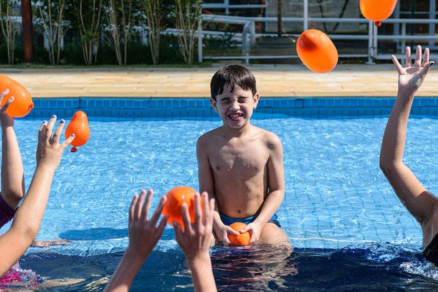 Criança de 8 anos sentada na beira da piscina e sendo alvo de várias bombas d'água de outras crianças.