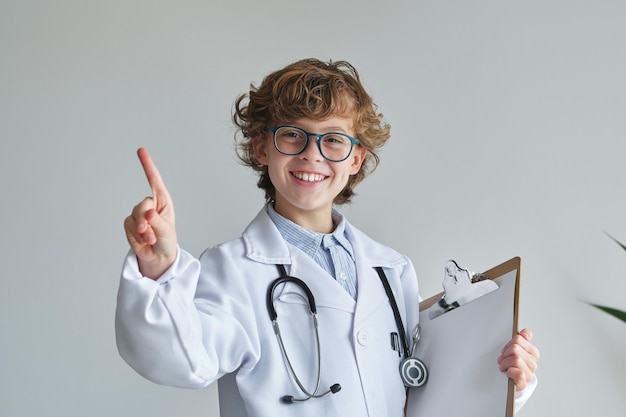 Criança de conteúdo com óculos e uniforme médico com estetoscópio e folha de papel mostrando o gesto da ideia enquanto olha para a câmera em fundo branco