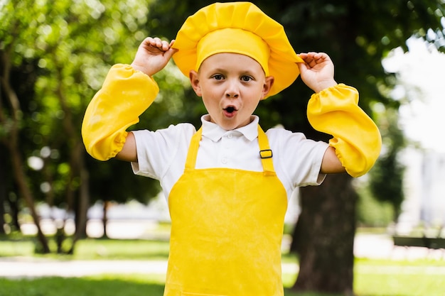 Criança de cozinheiro chocada com chapéu de chef amarelo e avental uniforme amarelo segurando chapéu de chef e surpresa ao ar livre