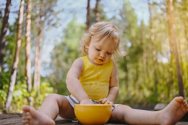 Criança de criança pequena engraçada loira bonita com roupas sujas e cara comendo comida de bebê ou