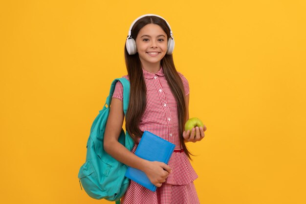 Criança de escola sorridente em fones de ouvido carrega mochila e pasta de trabalho com maçã para desintoxicação de almoço