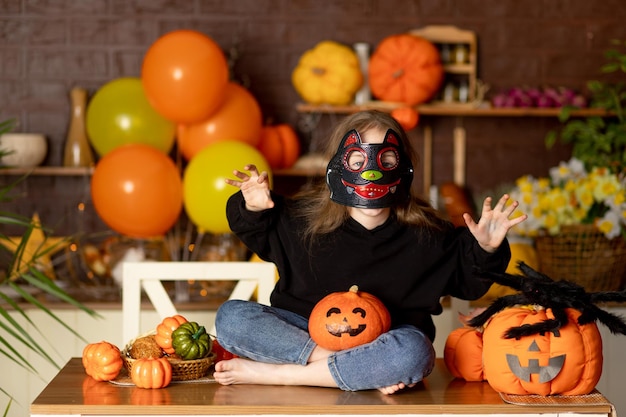 Foto criança de halloween uma garota com uma fantasia de bruxa e uma máscara de carnaval em uma cozinha escura com abóboras e uma grande aranha assusta com as mãos sorri se alegra