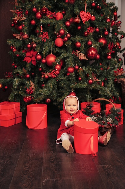 Criança de olhos azuis de um ano de idade com fantasia de Natal, brincando na sala de estar com caixas de árvore e presentes. Bebê fofo com emoção à noite de férias. Conceito de celebração familiar de Natal e Feliz Ano Novo